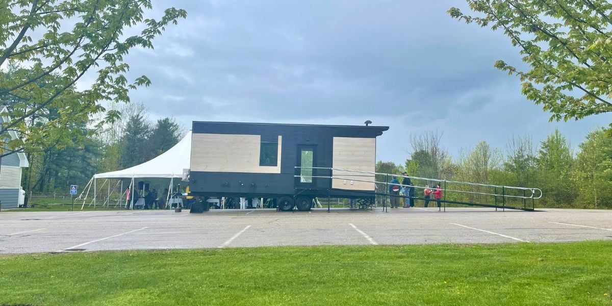 Wide view of the WheelPad® StudioPAD accessible modular home with an accessibility ramp beside a white event tent in a parking lot, surrounded by trees under cloudy skies.