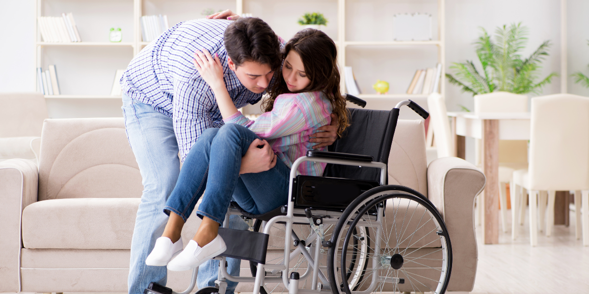 young male family caregiver helps a female into their wheelchair in their home 