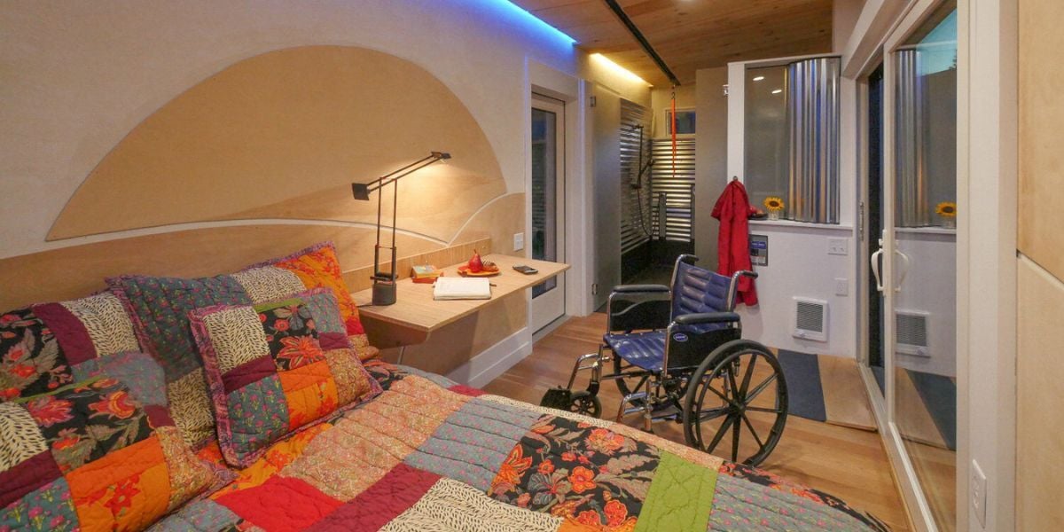 An interior photo of a staged SuitePAD, an accessible dwelling unit from WheelPad. A bed is in the forefront, with a wheelchair next to the bed. You can see deeper into the SuitePAD and see the main door, sliding doors, shower area, and desk included.