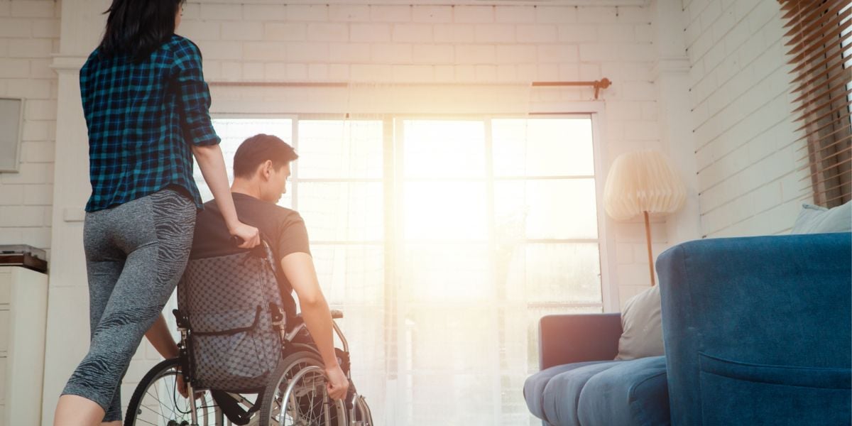 Female person helping push a man in a wheelchair in their home