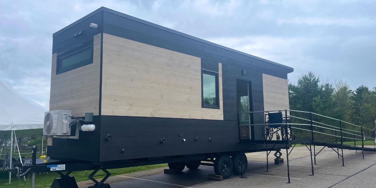 The WheelPad StudioPad, an accessible modular home, with a temporary accessibility ramp and wheelchair at the entrance, displayed outdoors at the Community Services of Addison County  offices in Middlebury, Vermont.